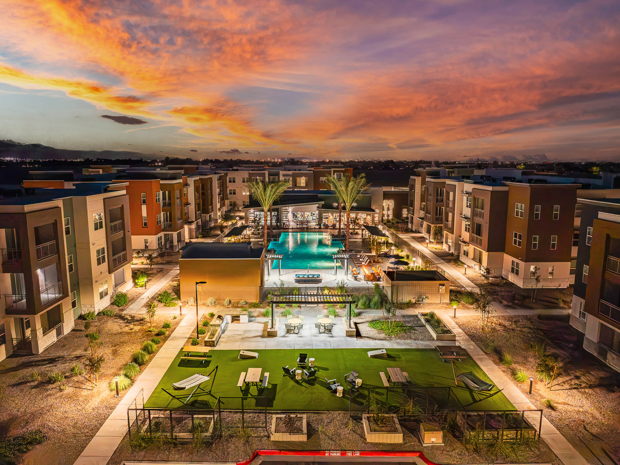 A modern apartment complex with a central pool and lounge area at sunset, surrounded by palm trees and gardens.