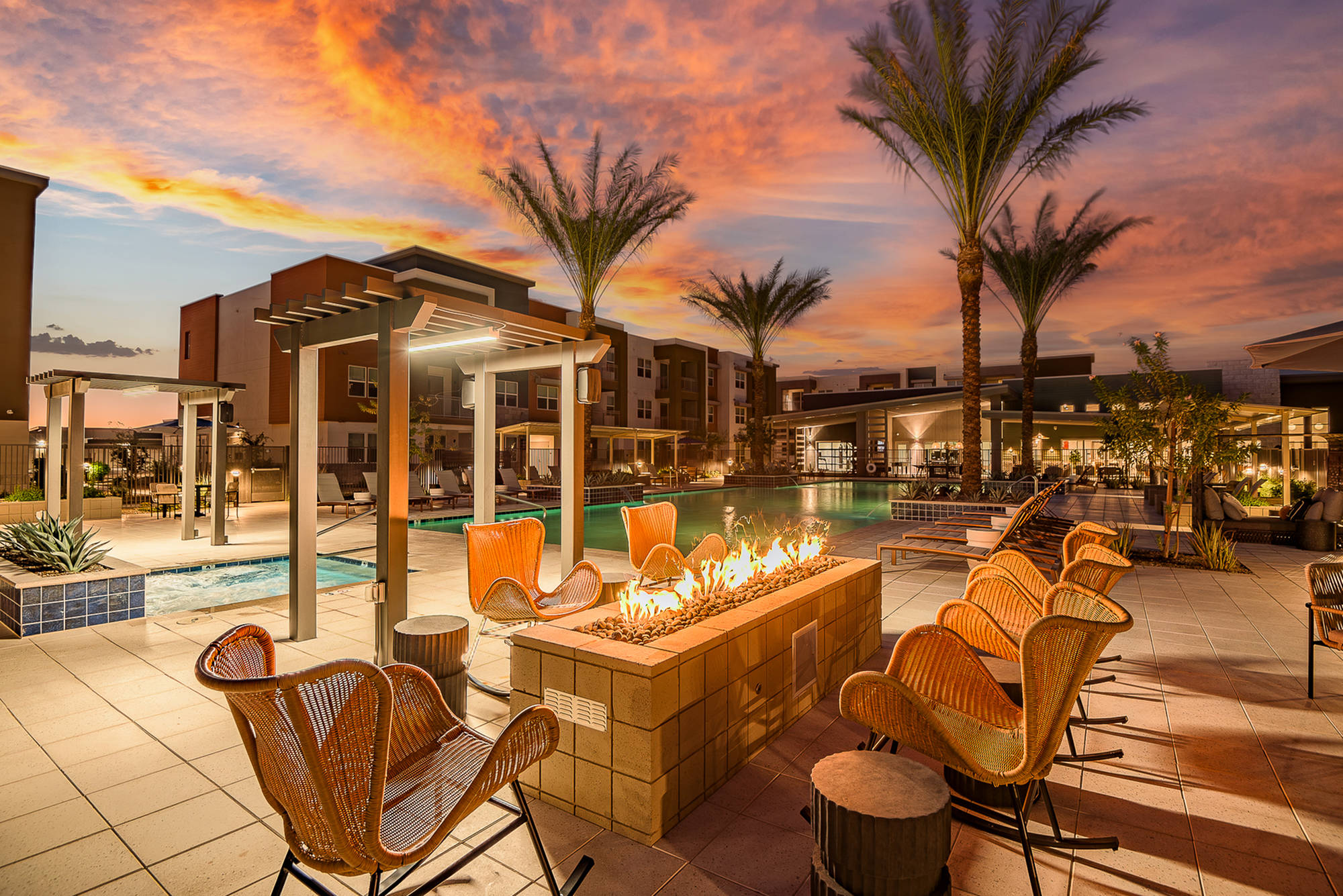 Outdoor pool area at sunset with palm trees, fire pit, lounge chairs, and modern apartment buildings.