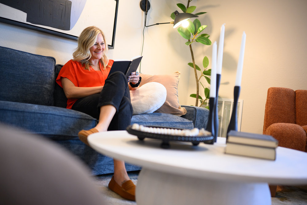 Woman in red top reading a book on a blue couch in a cozy, modern living room with a white coffee table.