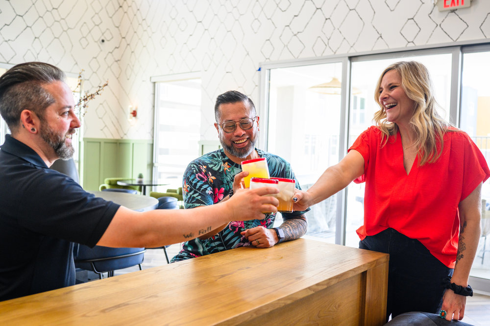 Three people smiling and clinking drinks together while standing around a wooden table in a bright room.