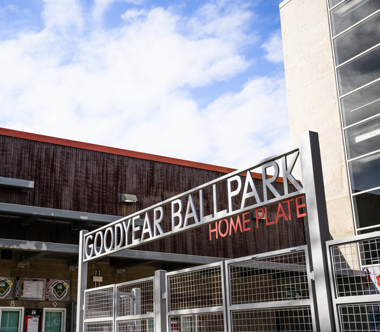Entrance sign for Goodyear Ballpark with "Home Plate" written below, under a partly cloudy sky.