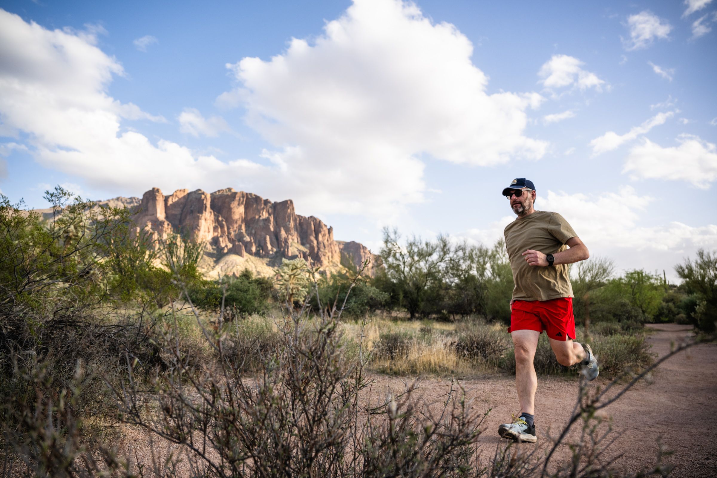 Man jogging on a desert trail with mountains and a partly cloudy sky in the background.