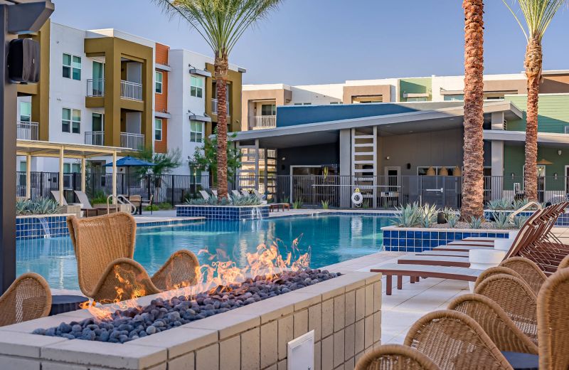 Modern apartment pool area with lounge chairs, fire pit, palm trees, and colorful buildings in the background.