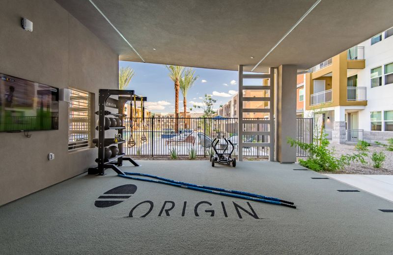 Outdoor gym area with equipment, TV, and "Origin" logo, overlooking a pool and apartment buildings.