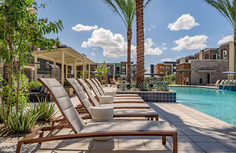 Lounge chairs beside a modern outdoor pool with palm trees and apartments in the background under a blue sky.