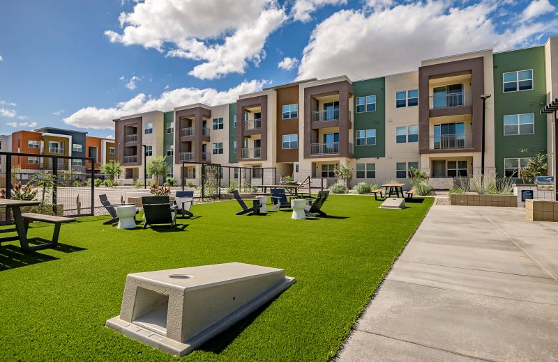 Modern apartment complex with three stories, outdoor picnic tables, and a cornhole game area on green lawn.