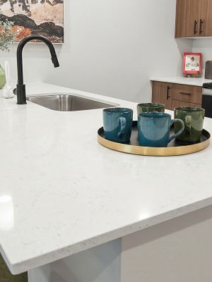 Modern kitchen with a white quartz island, green chairs, a sink, and mugs on a tray.