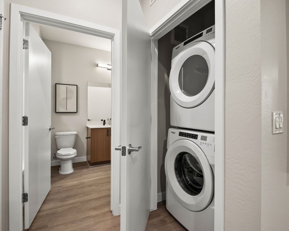 Stacked washer and dryer in a closet next to a bathroom with wood floor and modern decor.