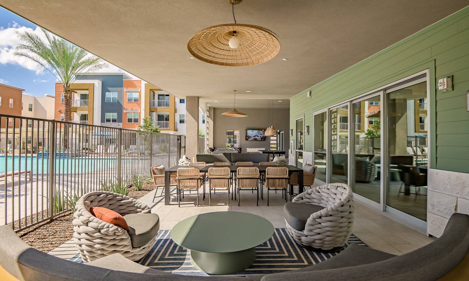 Modern outdoor lounge with woven chairs, dining table, TV, and view of a swimming pool and apartment buildings.
