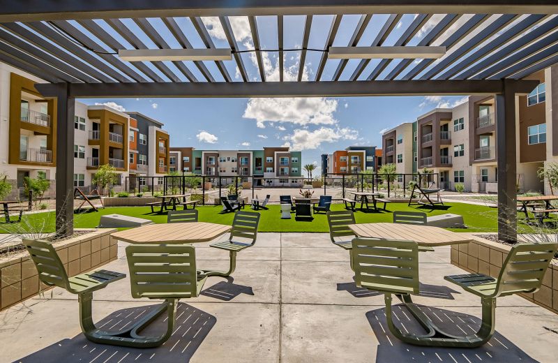 Outdoor seating area under pergola with tables and chairs, facing modern apartment buildings and green lawn.